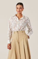 A woman is wearing a long-sleeved, white lace button-up shirt with a floral pattern and a classic collar. She has paired it with high-waisted, wide-leg beige trousers that feature pleats and a belt loop.